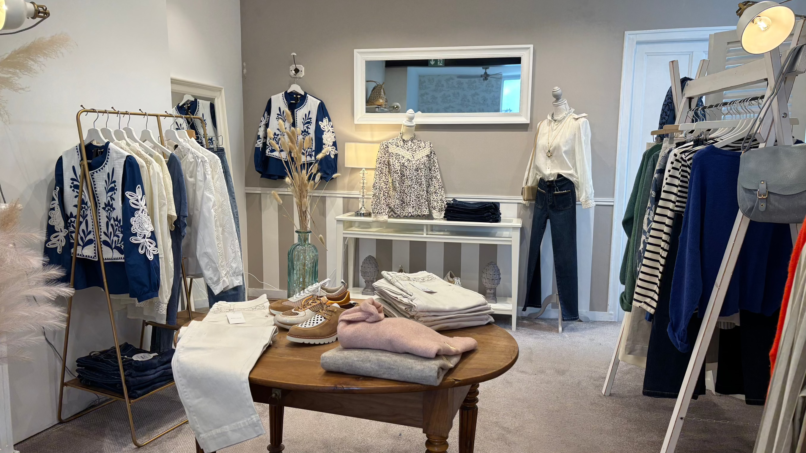 Un magasin de vêtements avec des articles bien organisés. On y voit des manteaux, des chemisiers blancs et bleus ornés de broderies, des pantalons blancs et des chaussures disposés sur une table en bois. Des étagères et des porte-vêtements sont également présents, offrant une vue d'ensemble de la boutique dans des tons neutres et lumineux.